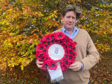 Alex stands in front of autumn foliage holding a wreath of poppies. In the centre of the wreath is the symbol of the Scottish Parliament.